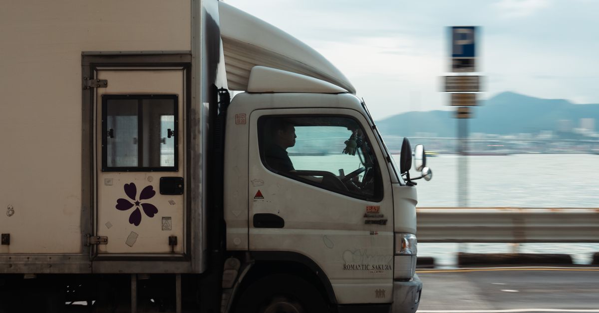 A delivery truck driving along a coastal road with a mountain backdrop on a clear day.